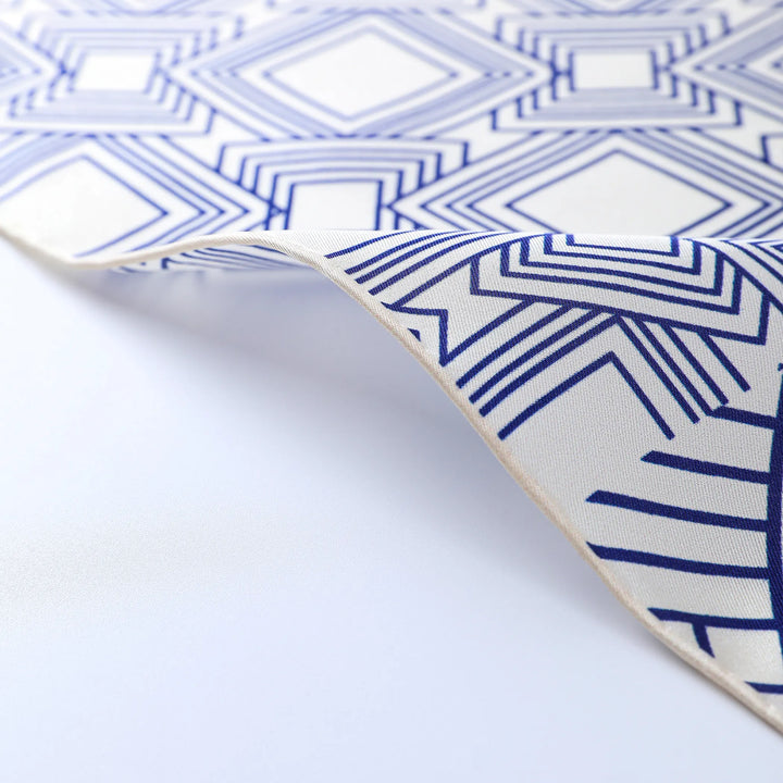 A close-up view of the Land Off Blue Square Silk Scarf from SusanSilk displays a blue and white geometric pattern featuring diamond and linear designs. The scarf is slightly curled at the edge.