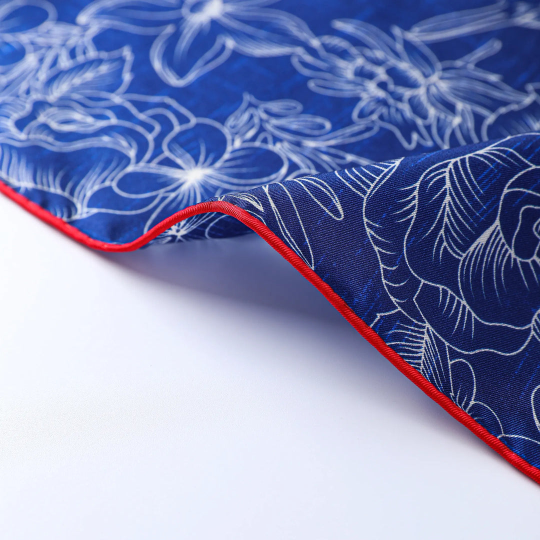 Close-up of the Deep Blue Square Silk Scarf by SusanSilk, showcasing its blue silk fabric adorned with white floral patterns and a red edge. One corner of the scarf is slightly lifted, revealing its underside.