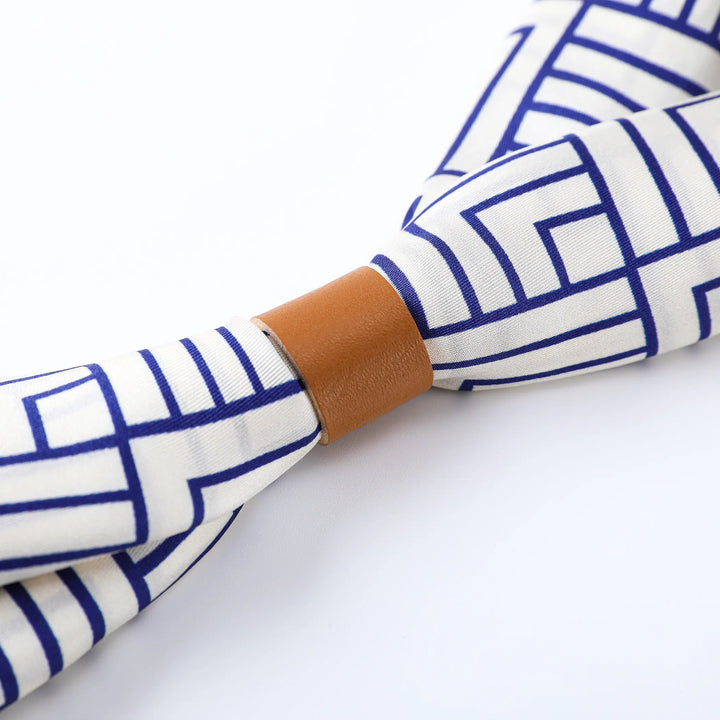 Close-up of the Land Off Blue Square Silk Scarf by SusanSilk, featuring intersecting blue lines on a white background, cinched with a brown leather band.