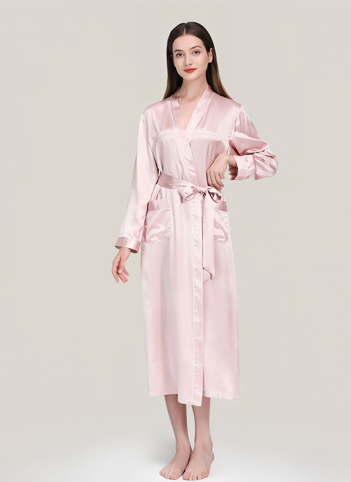 A person wearing a SusanSilk 22 Momme Contrast Piping Long Silk Robe in light pink, standing barefoot against a plain light background.