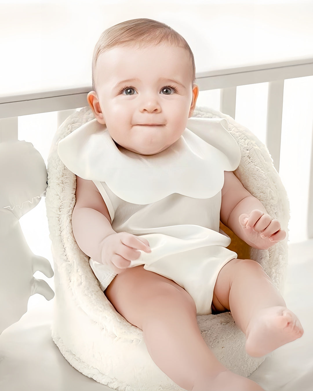 A baby with light brown hair sits in a soft, white cushioned seat. The baby wears a white outfit adorned with the SusanSilk 19 Momme Baby Silk Petal Bib, featuring a large ruffled collar made of delicate silk, and looks up with a slight smile.