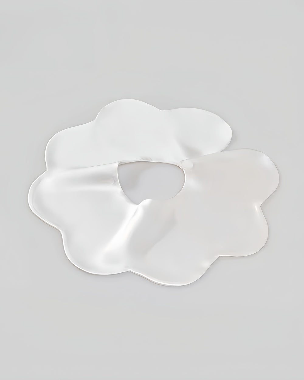 A 19 Momme Baby Silk Petal Bib by SusanSilk, featuring a white, flower-shaped design with irregular contours and a central hole, reminiscent of delicate silk, set against a plain light grey background.