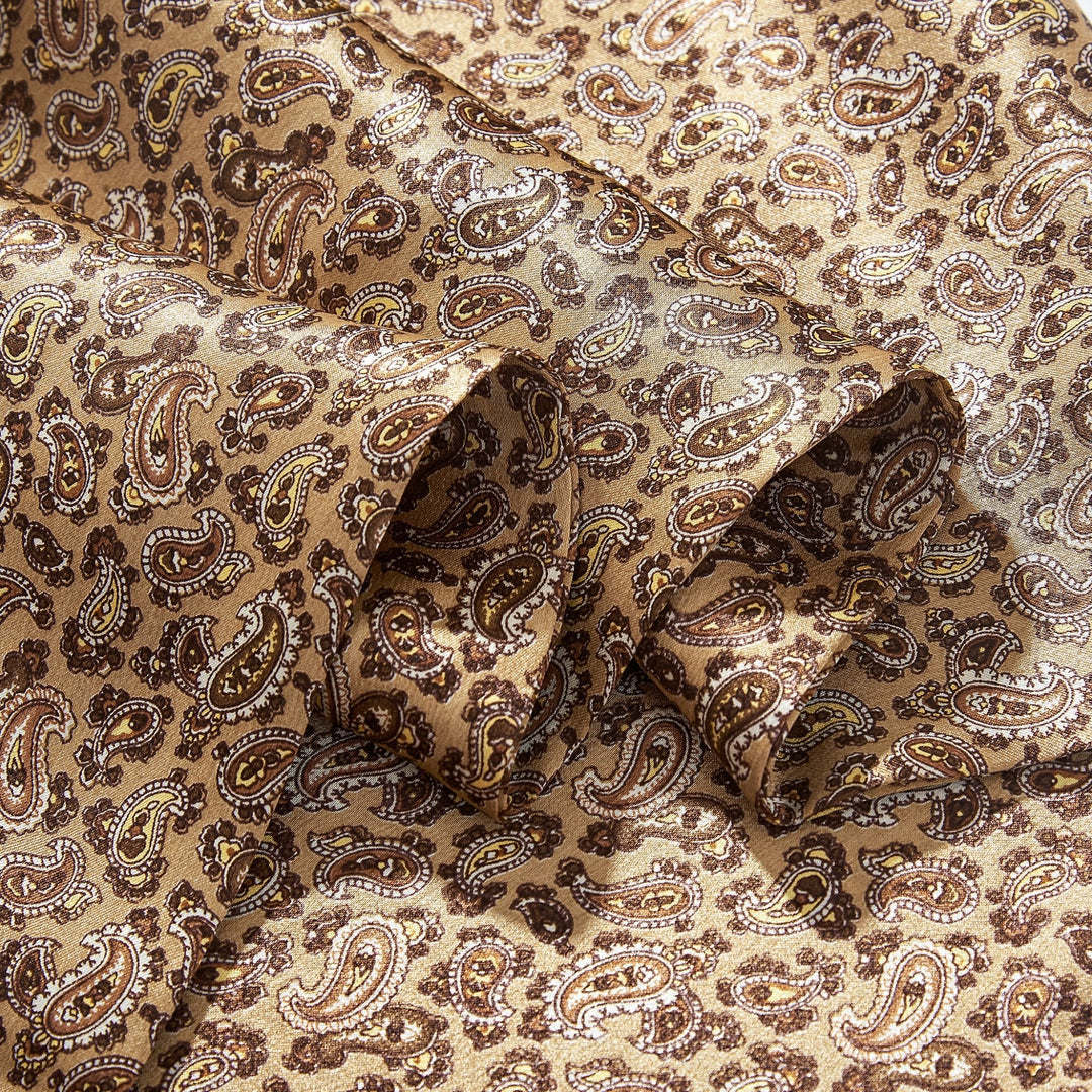 A close-up of a folded fabric from the SusanSilk Paisley Series Long Silk Scarf, showcasing a golden brown and beige paisley pattern.