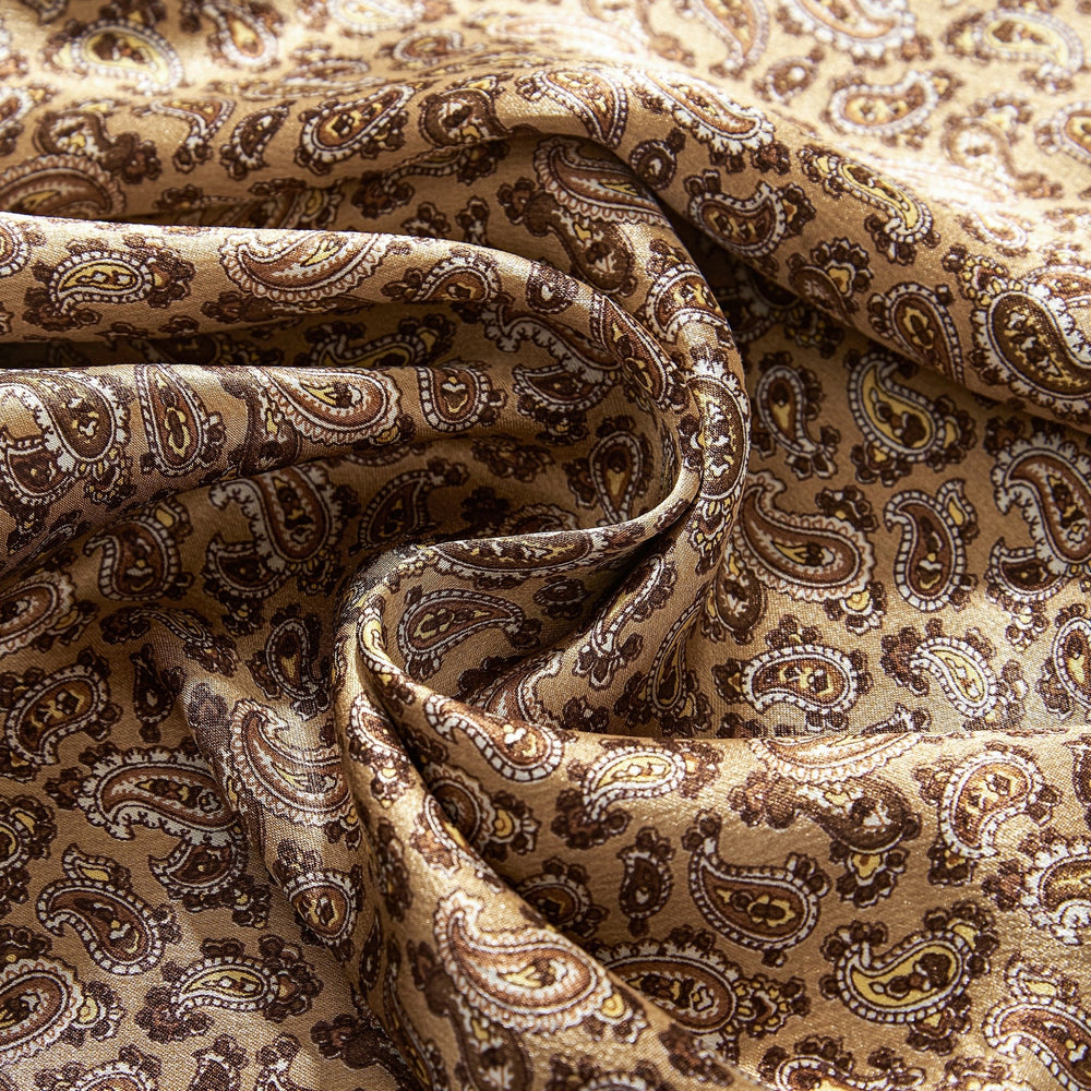 A close-up of the Paisley Series Long Silk Scarf from SusanSilk, featuring a beige fabric adorned with an intricate paisley pattern in shades of brown and yellow, showcasing smooth folds and detailed designs reminiscent of luxurious craftsmanship.