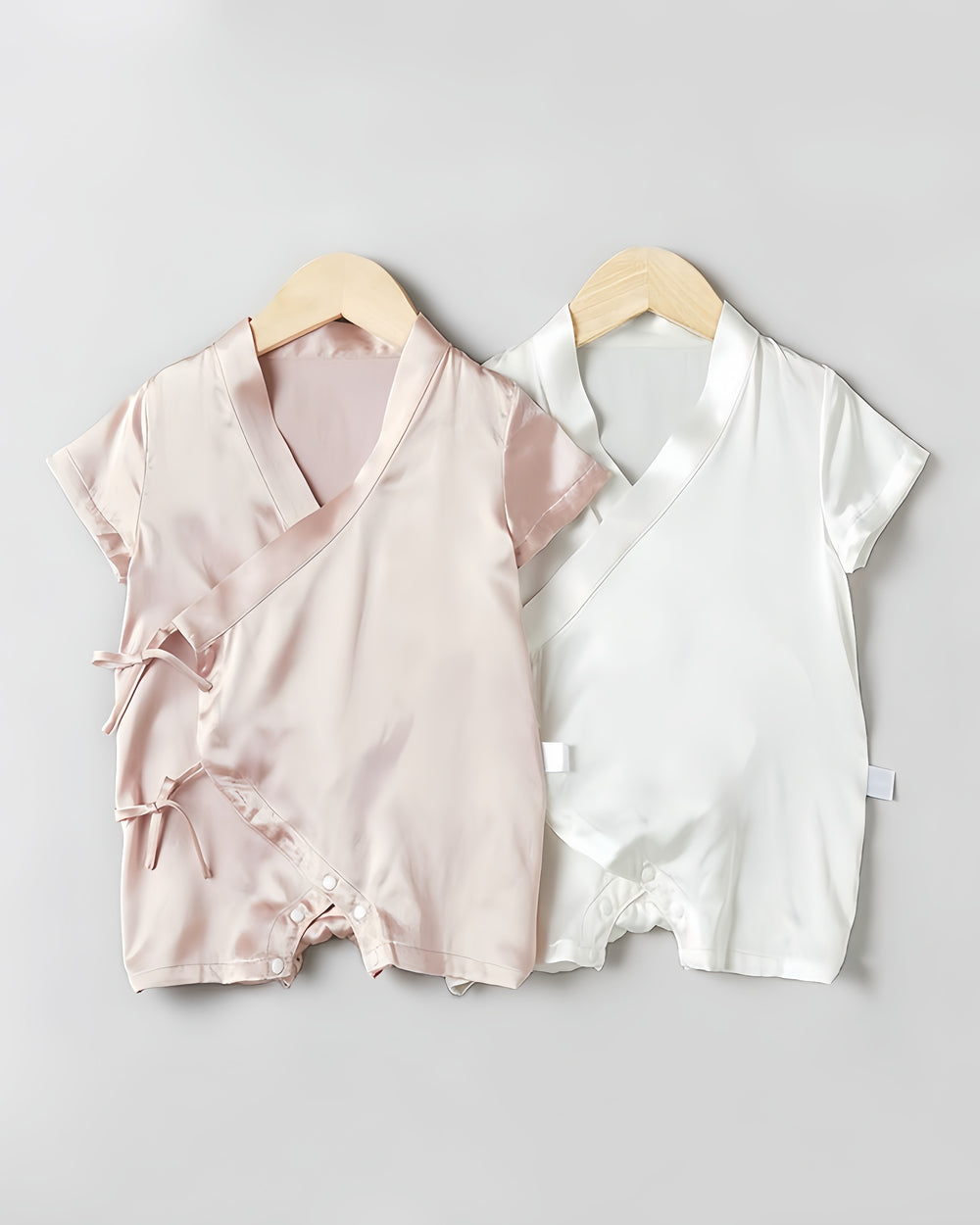 Two 19 Momme Silk Baby Button Lace-Up Onesies by SusanSilk, one in light pink and one in white, made of soft silk, hang on wooden hangers against a plain background. Each features side ties and a snap closure at the bottom.