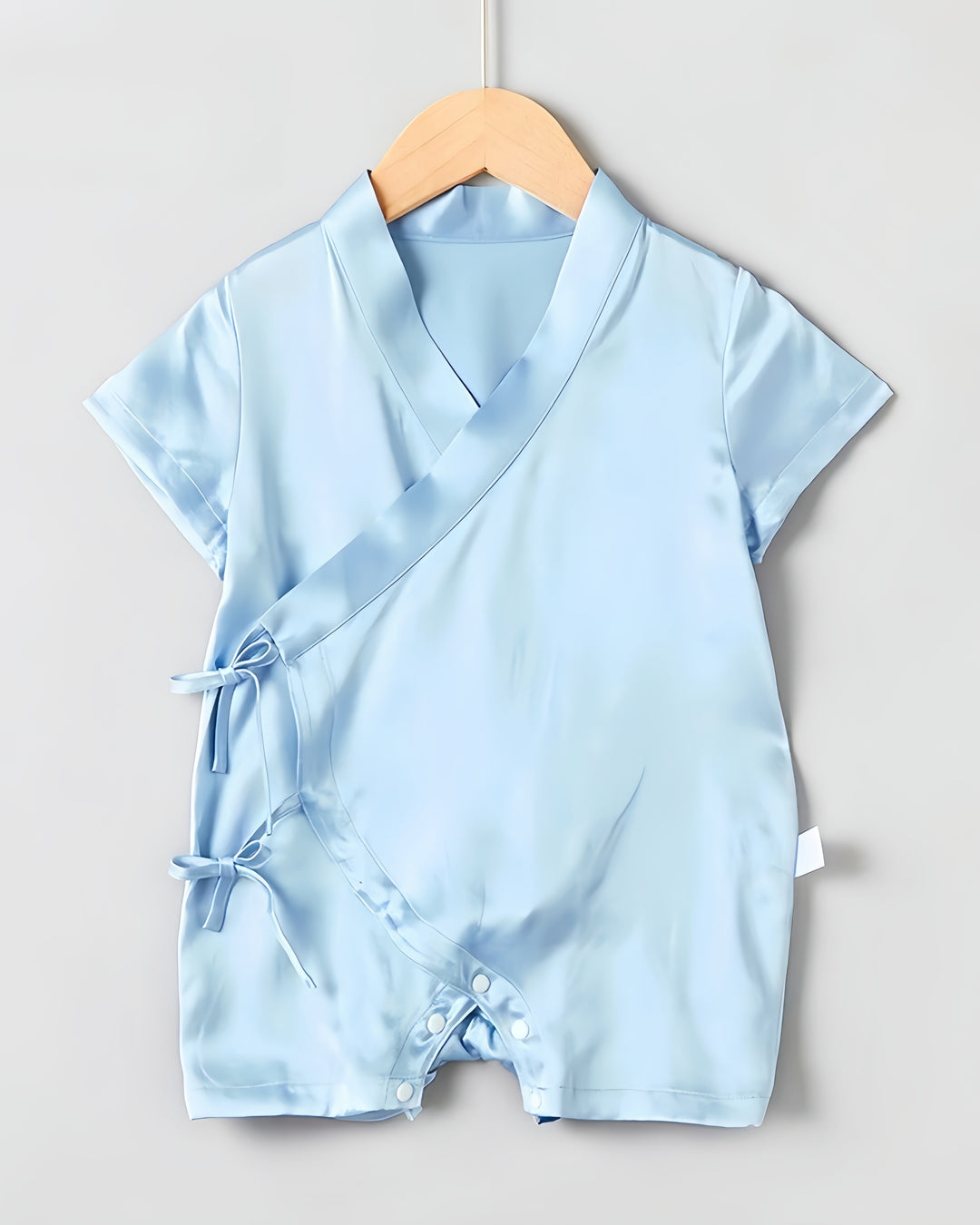 Introducing the 19 Momme Silk Baby Button Lace-Up Onesie by SusanSilk: a light blue satin romper showcasing short sleeves and a luxurious wrap-style front. This exquisite piece features two side tie closures and convenient snap buttons at the bottom, elegantly displayed on a wooden hanger against a plain background.