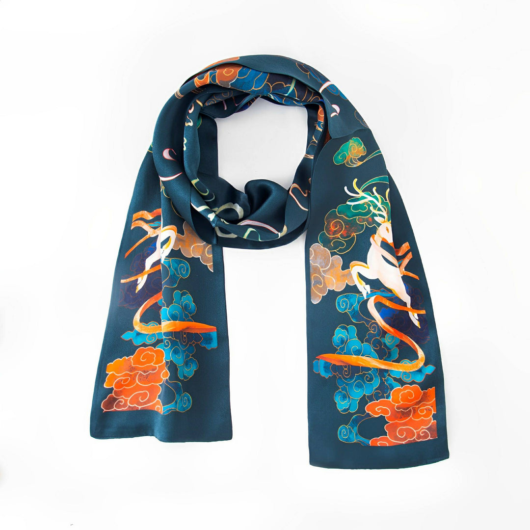 A Fairy Deer Long Mulberry Silk Scarf from SusanSilk is laid flat, showcasing colorful illustrations of a white mythical creature surrounded by orange, blue, and green clouds against a dark blue background.