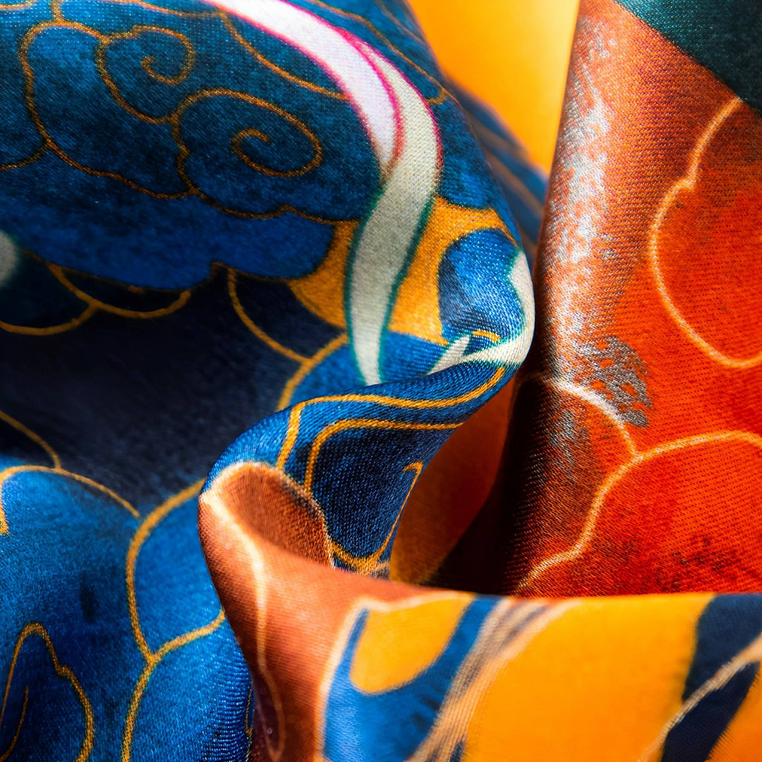 Close-up of the Fairy Deer Long Mulberry Silk Scarf by SusanSilk, showcasing its vividly colored fabric adorned with intricate blue, orange, and gold patterns. The material appears to be folded or draped, highlighting its smooth texture and detailed design.