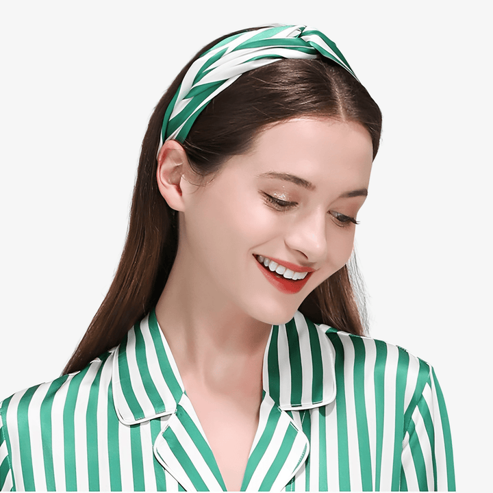 A woman with long brown hair, adorned in a green-and-white striped shirt and a matching Stripe Criss Cross Silk Headband by SusanSilk, smiles softly while looking down. The satin sheen of her outfit hints at its luxurious silk material.