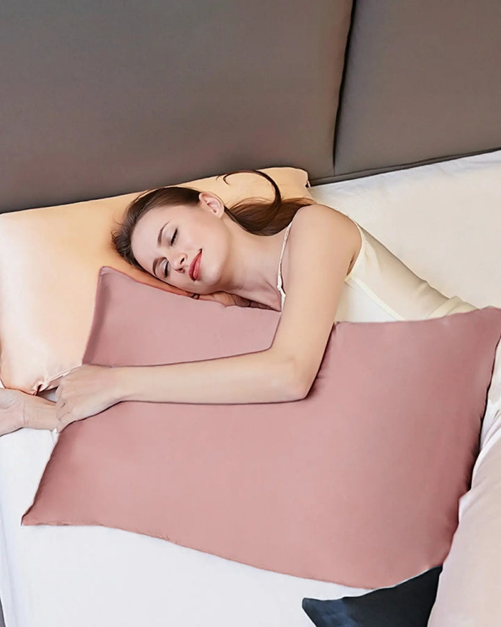 A person lies on a bed with their head on a cream-colored pillow, embracing a large pink silk pillow from the **New Arrival 25 Momme Beauty Silk Pillowcase Zipper** collection by **SusanSilk** while sleeping.