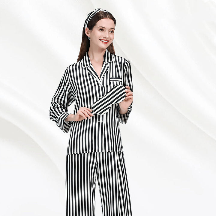 A woman wearing the 19 Momme Chic Stripe Silk Pajamas Set by SusanSilk, along with a matching headband, stands against a white background, holding a striped silk sleep mask.