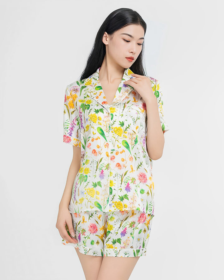 A woman models the Floral Short Silk Pajamas Set by SusanSilk, showcasing its luxurious design. The set features short sleeves and shorts adorned with a vibrant array of colorful flowers and leaves against a pristine white background.