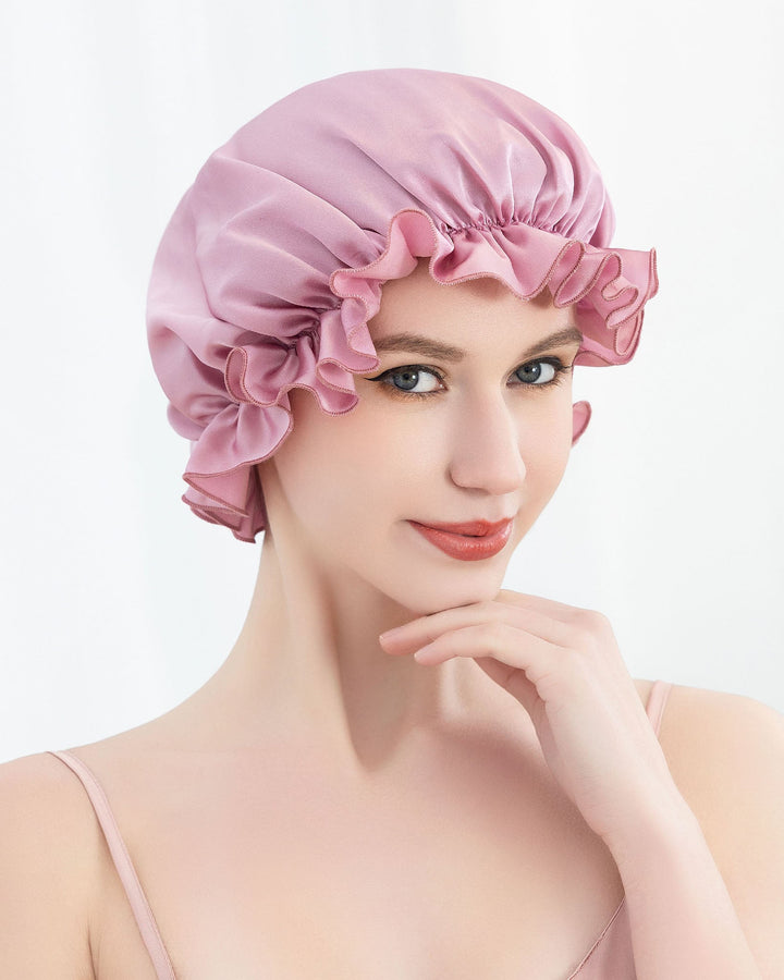 A woman with fair skin wearing the SusanSilk Flounced Silk Bonnet For Sleeping and a light pink camisole is looking at the camera with her hand resting under her chin.