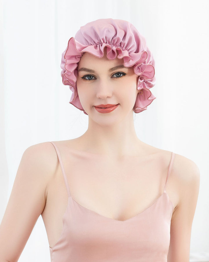 A person wearing a flounced pink silk bonnet from SusanSilk and a matching satin camisole, standing against a white background.