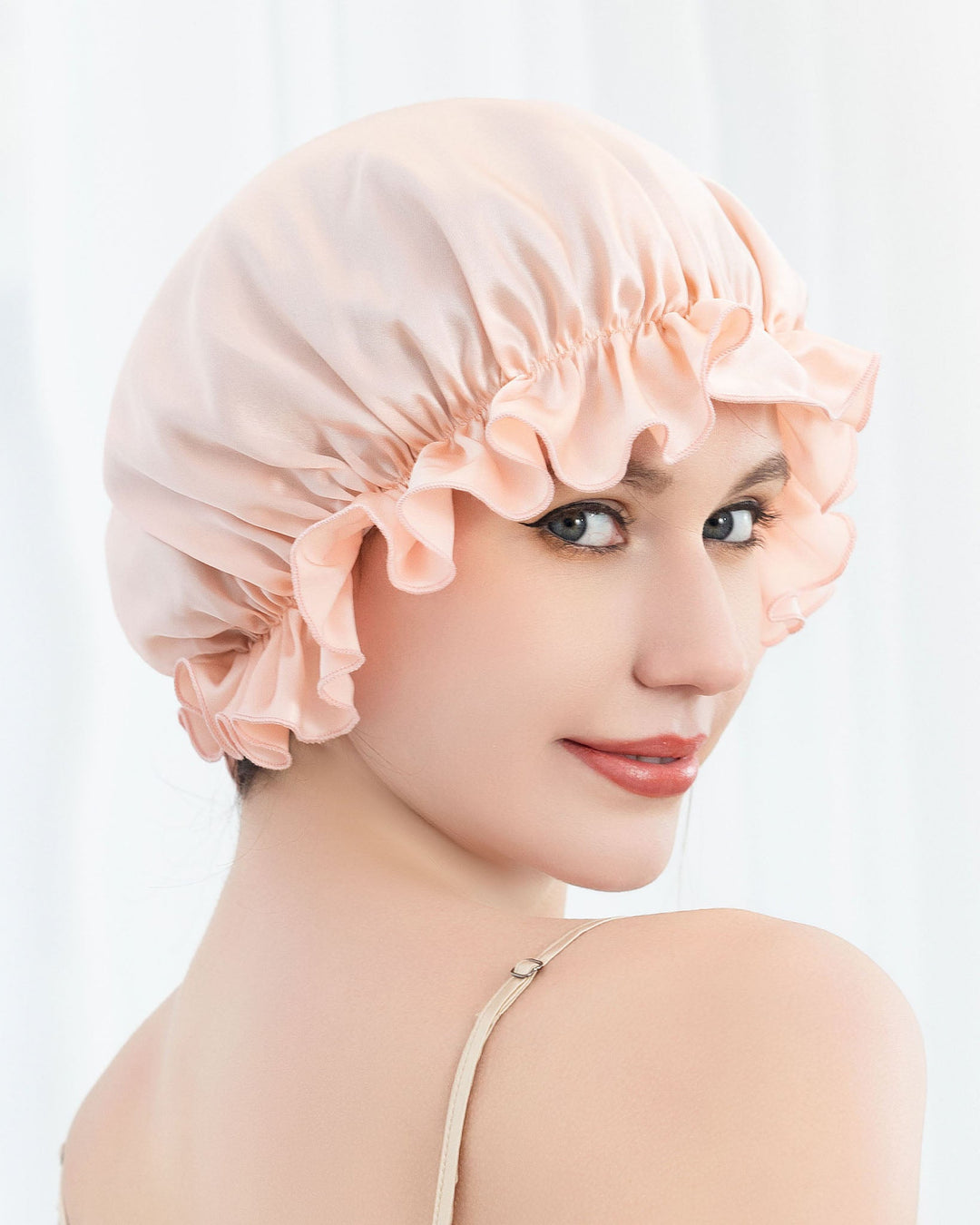 A person is shown wearing the SusanSilk Flounced Silk Bonnet For Sleeping in a peach color, with ruffled edges, looking over their shoulder with a slight smile.