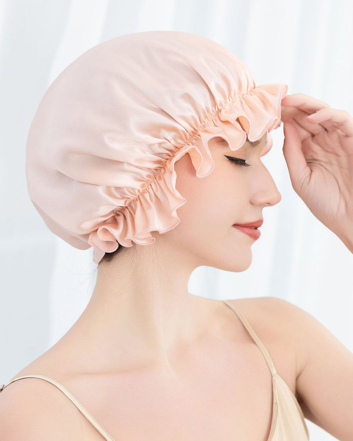 A person wearing a peach-colored SusanSilk Flounced Silk Bonnet For Sleeping is gently touching the edge with their fingertips while gazing downward.