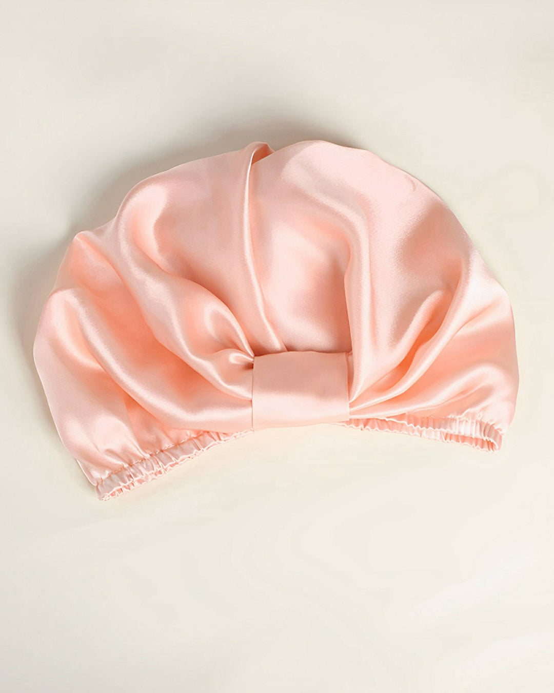 An Elegant Pleated Silk Bonnet For Sleeping by SusanSilk, featuring a light pink satin finish, an elastic band, and a central fabric fold for ultimate hair protection.