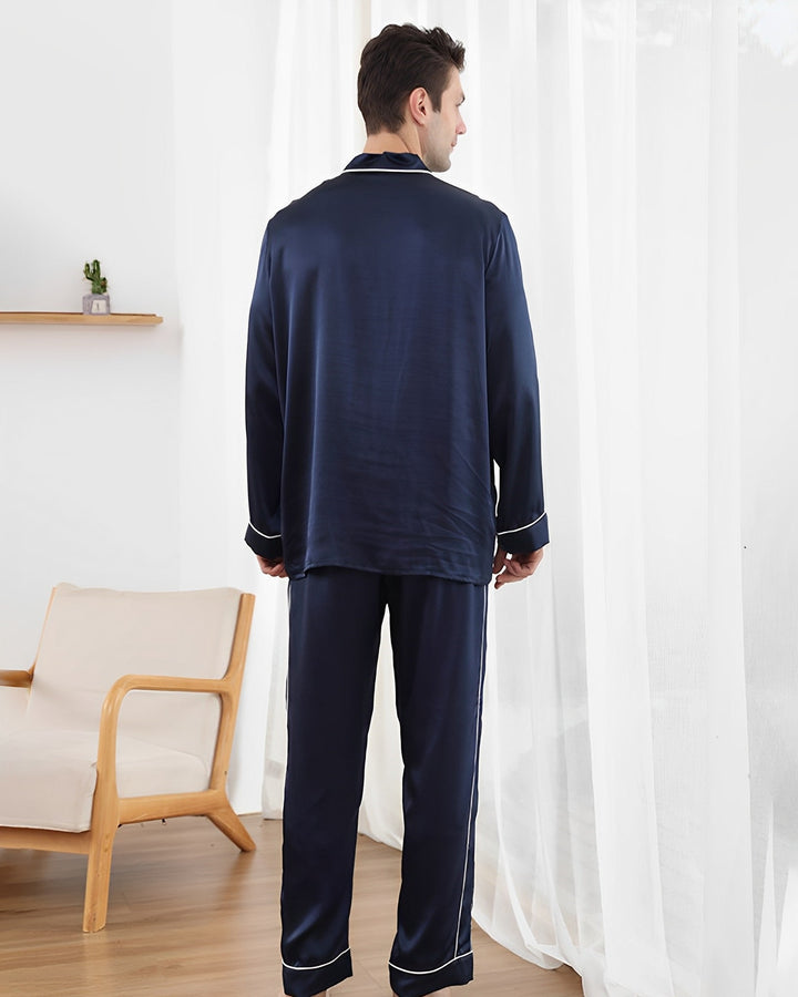 A man wearing the 19 Momme Eton Collar Silk Pajamas Set from SusanSilk stands with his back to the camera in a room with light curtains and wooden furniture.