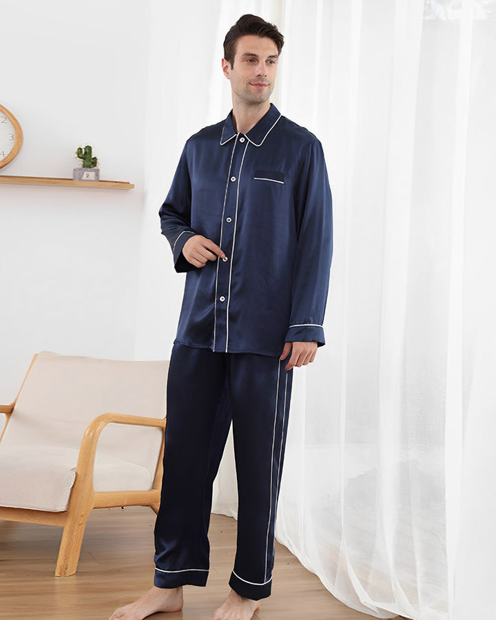 A man stands in a room wearing the SusanSilk 19 Momme Eton Collar Silk Pajamas Set in dark blue with white piping. He is barefoot on a wooden floor, beside a cream-colored chair with a wooden frame and next to a sheer curtained window.