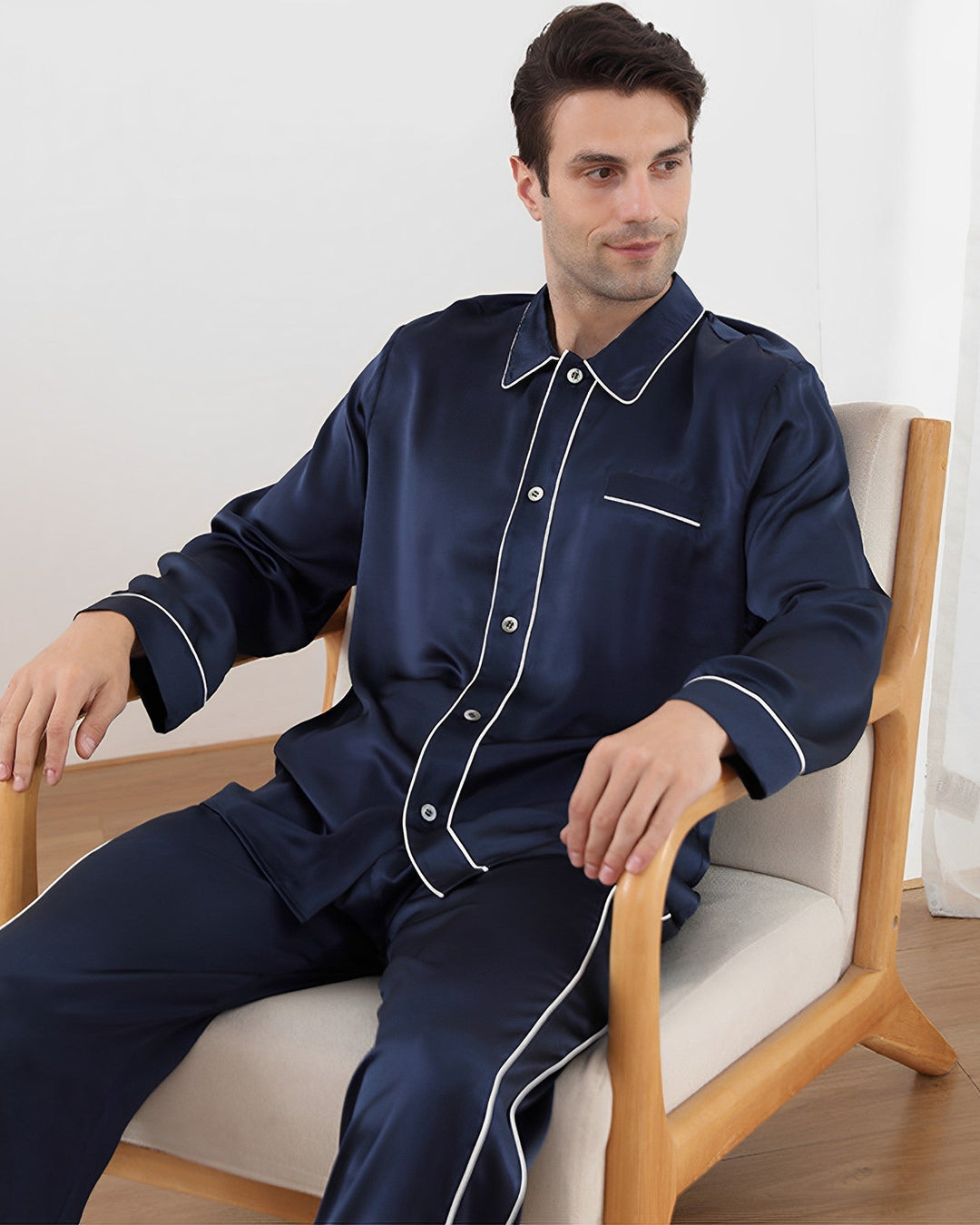 A man sits on a wooden armchair wearing the 19 Momme Eton Collar Silk Pajamas Set from SusanSilk, featuring navy blue silk with white piping.