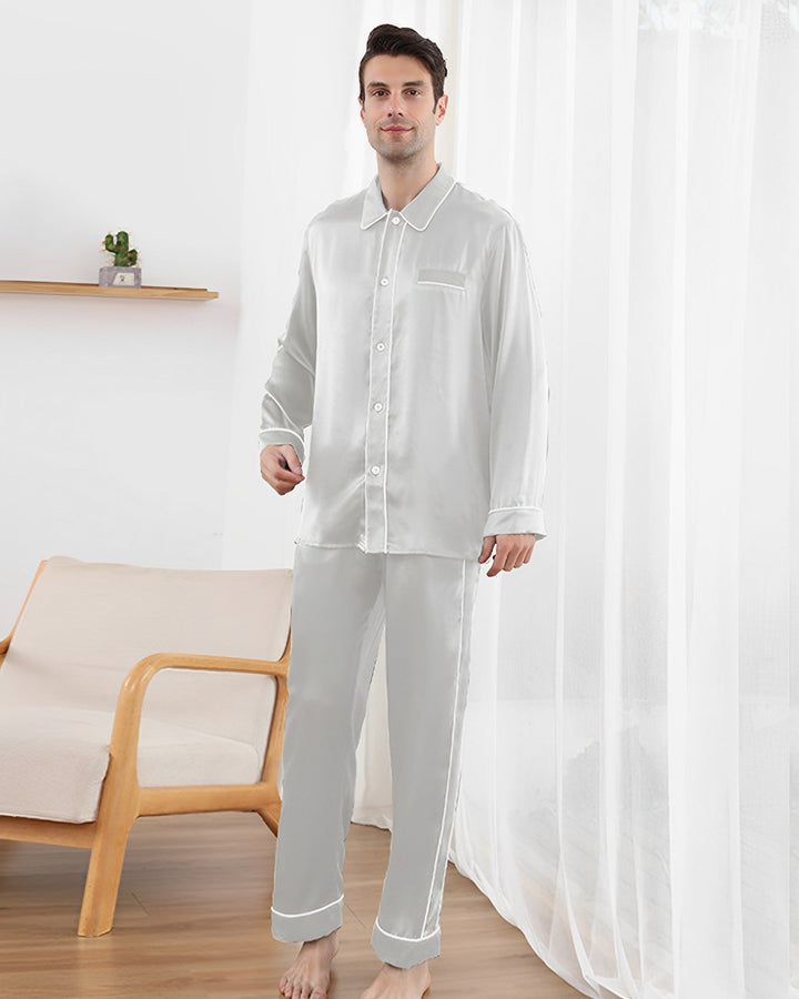 A man stands in a room wearing the 19 Momme Eton Collar Silk Pajamas Set from SusanSilk, featuring a light grey silk button-up shirt. A beige chair and a small wooden shelf adorned with plants add to the decor, while sheer white curtains partially reveal a window.