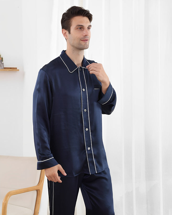 A man wearing a SusanSilk 19 Momme Eton Collar Silk Pajama Set with white piping stands in a room with a chair and white curtains.