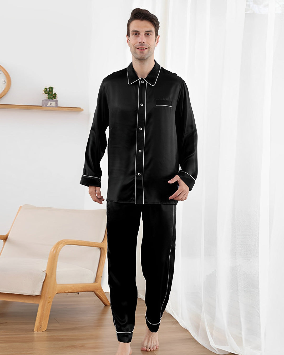 A man is standing in a living room wearing a SusanSilk 19 Momme Eton Collar Silk Pajamas Set with white piping, next to a wooden chair and a white curtain.