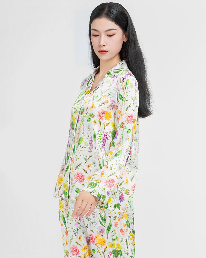 A woman with black hair stands against a plain backdrop, wearing the 19 Momme Floral Silk Pajama Set by SusanSilk. She looks downwards with a neutral expression.