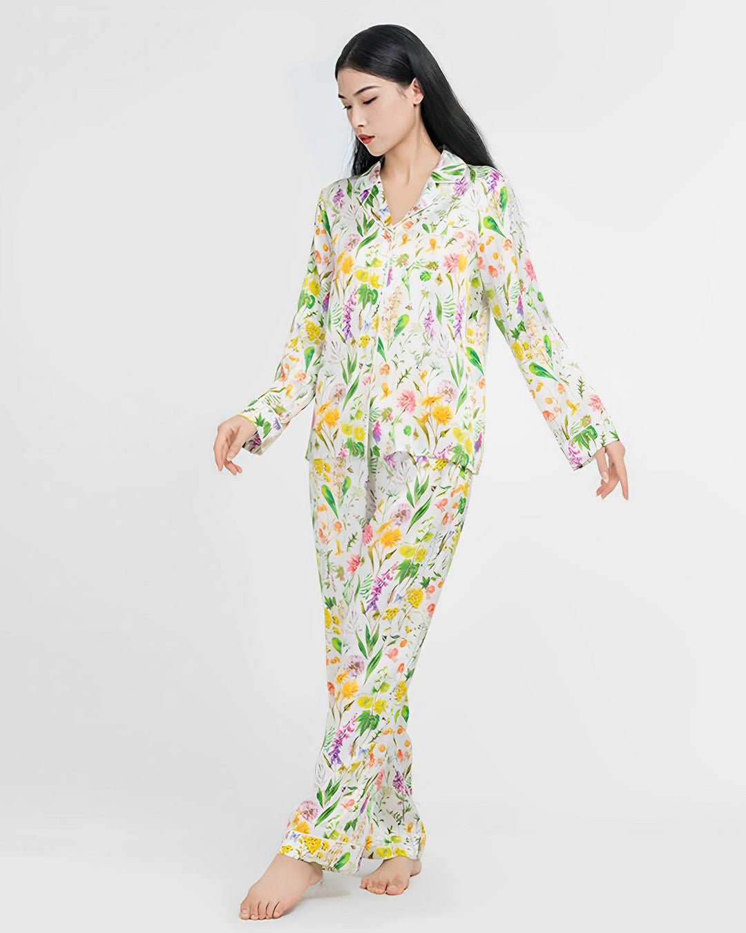 A woman stands against a white background wearing the 19 Momme Floral Silk Pajamas Set from SusanSilk, made from luxurious silk and featuring a colorful floral pattern. She is barefoot and looking downward.