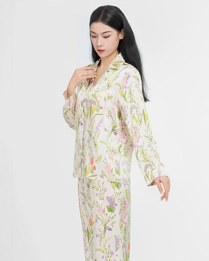 A woman with long black hair is wearing a 19 Momme Floral Silk Pajamas Set by SusanSilk. She is looking down with one hand near her collar and the other extended downward.