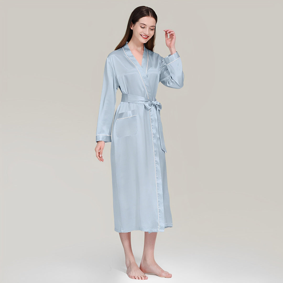 A woman stands smiling, barefoot, and gracefully adorned in a 19 Momme Contrast Piping Long Silk Robe by SusanSilk. This elegant attire features a belt and two pockets, perfectly accentuated against the plain, light-colored background.