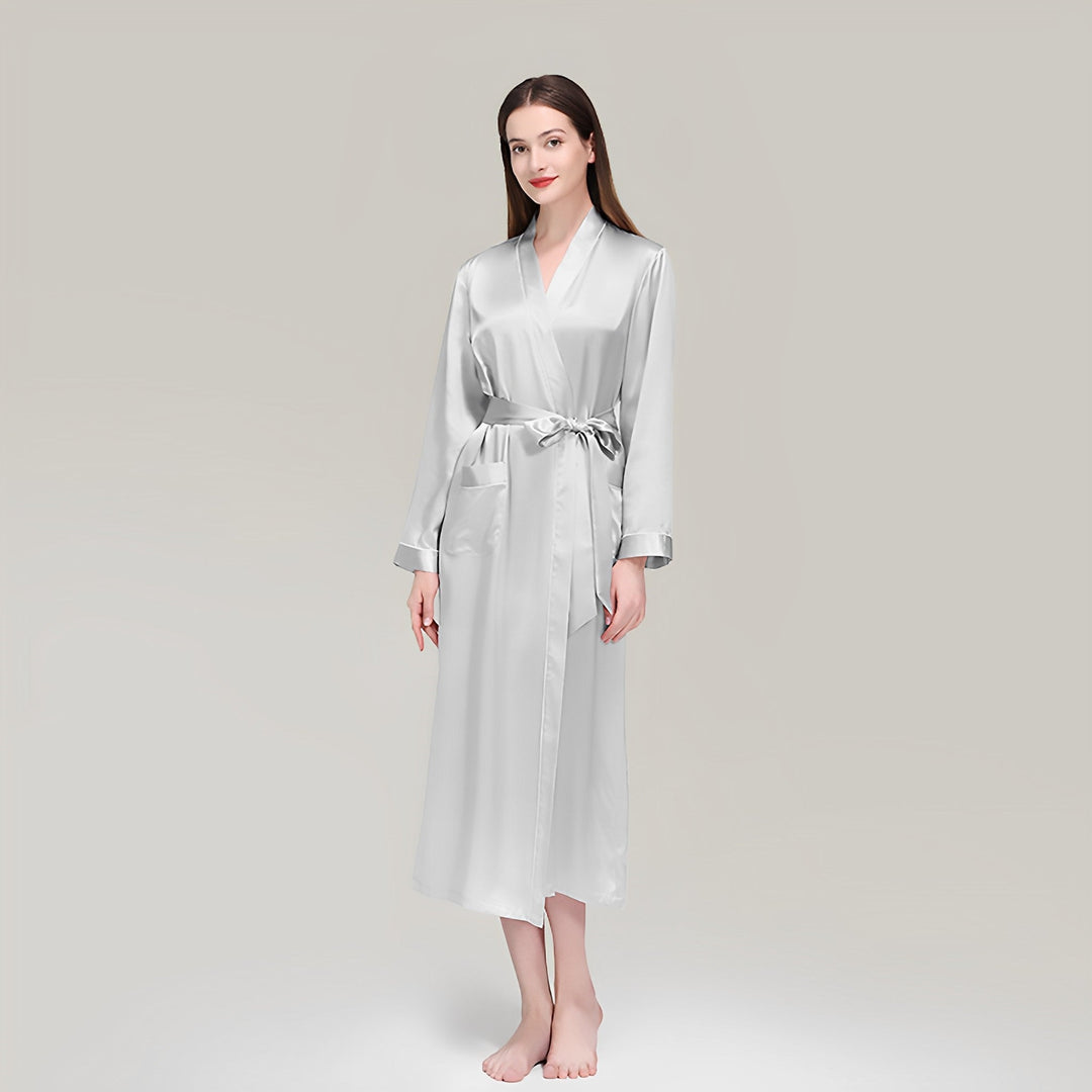 A woman stands barefoot and smiling slightly, wearing the SusanSilk 19 Momme Contrast Piping Long Silk Robe for women. The long, silver silk robe with a tied belt contrasts elegantly against the plain, light gray background.