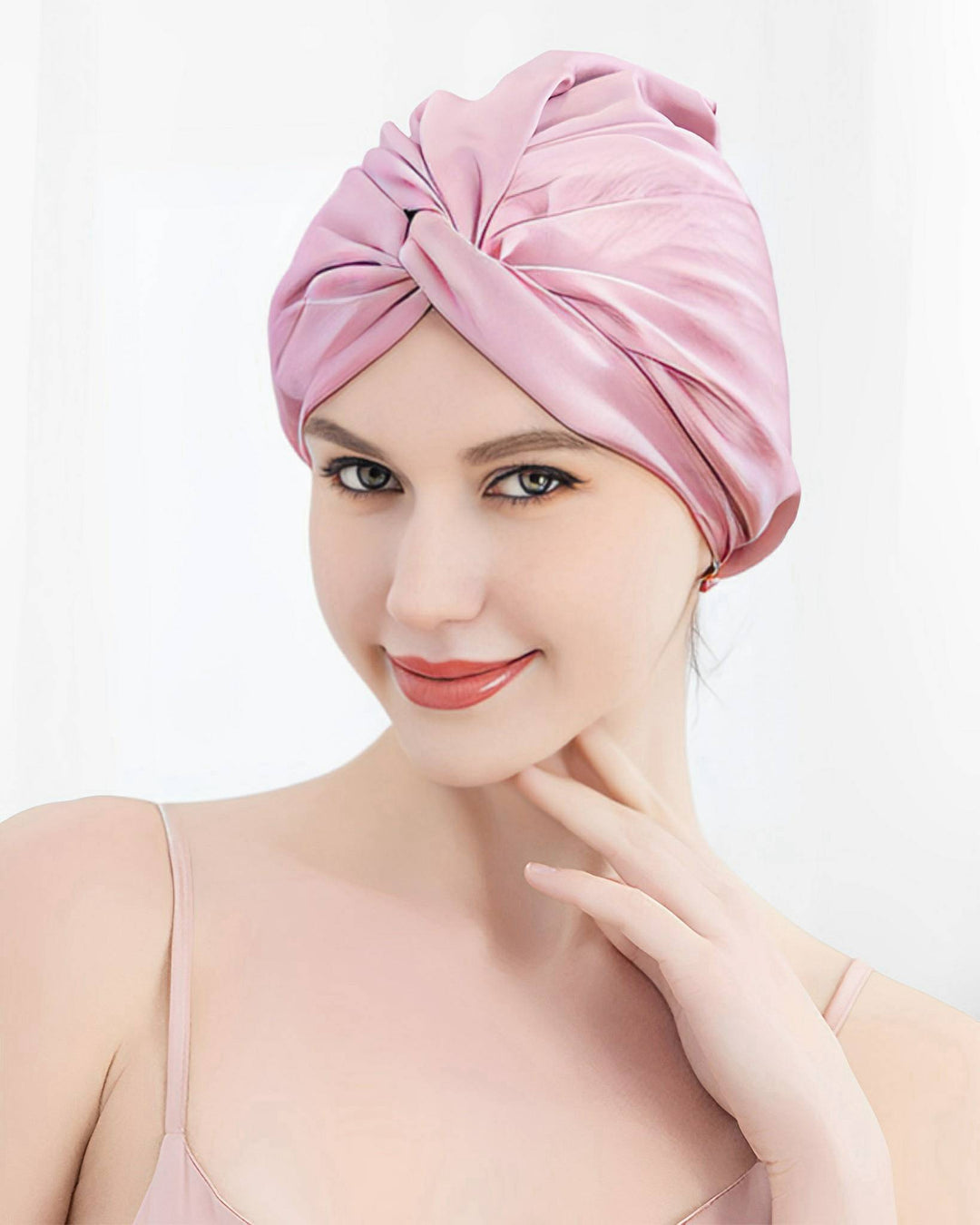 A woman sporting a SusanSilk Luxury Turban Silk Sleep Cap and an elegantly matching silk dress, smiling softly while touching her chin with one hand.