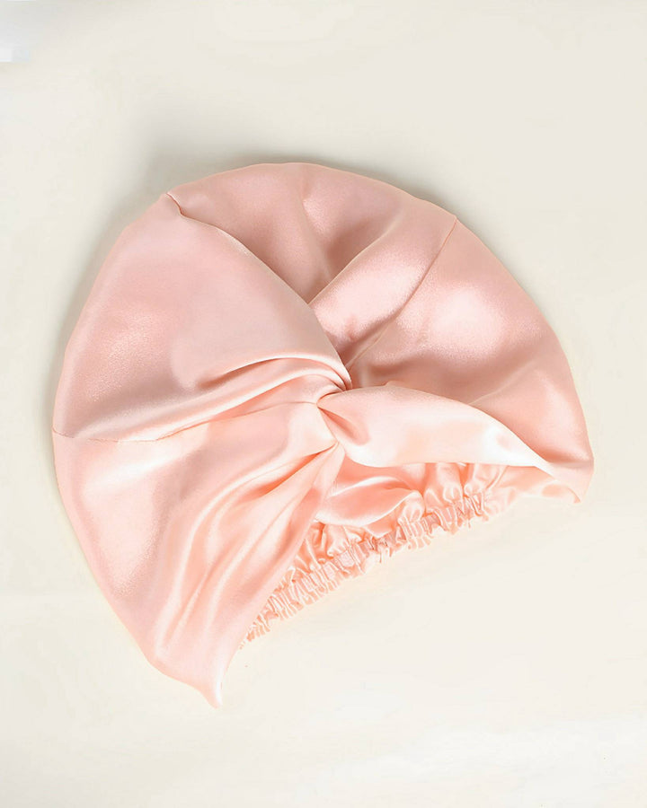 A SusanSilk Luxury Turban Silk Sleep Cap in pink satin, featuring a gathered elastic band at the edge, placed on a light-colored surface.