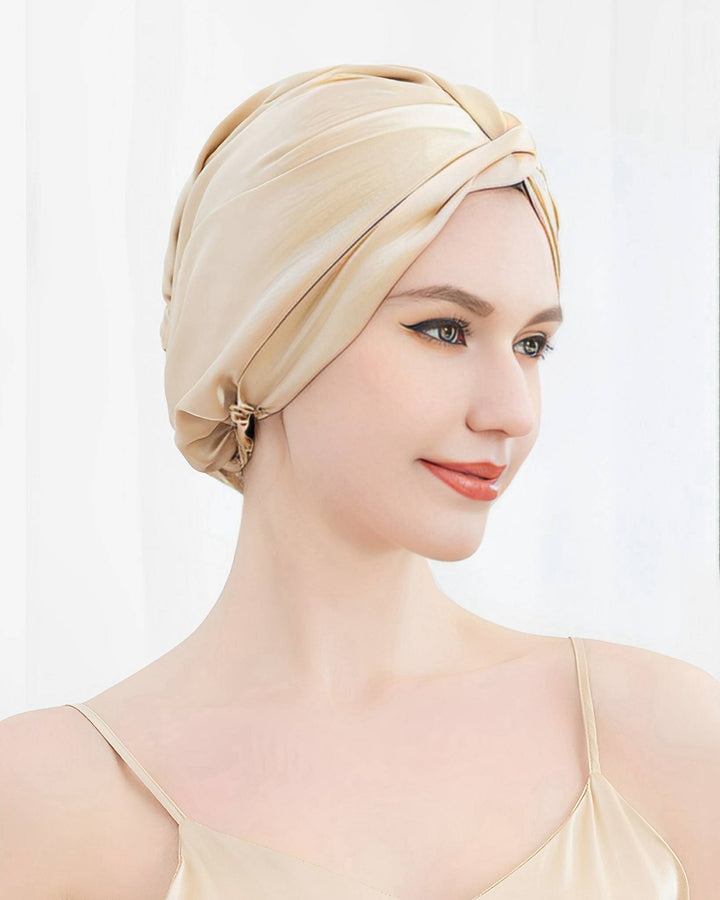 A woman with fair skin and light brown hair is wearing a beige Luxury Turban Silk Sleep Cap by SusanSilk and a matching SusanSilk spaghetti strap top, smiling slightly, against a white background.