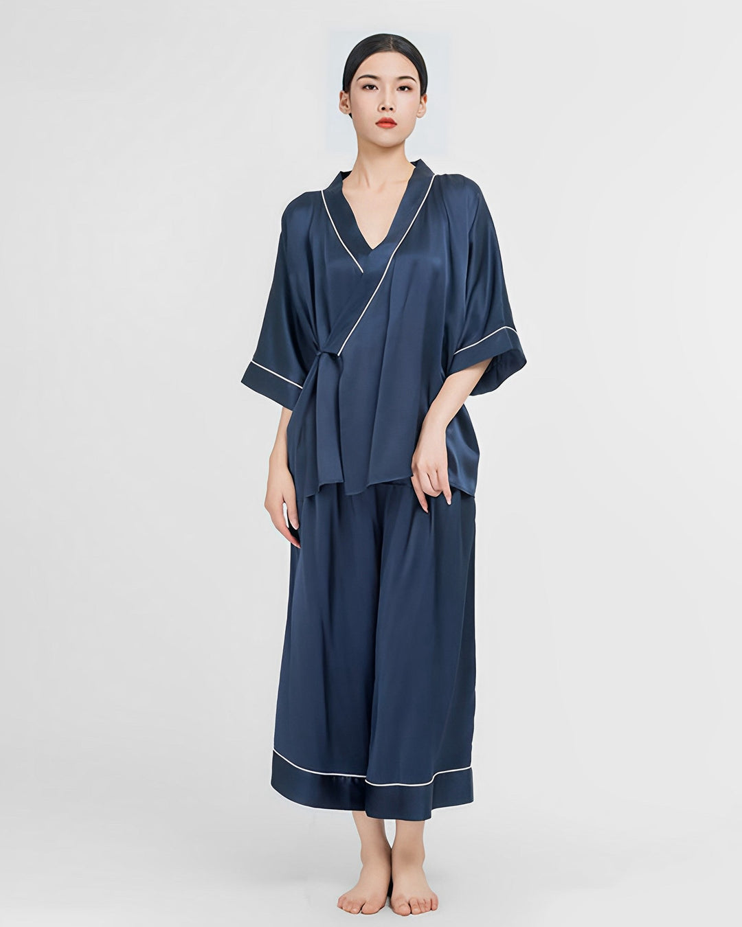 A person stands against a plain background wearing the stunning 19 Momme Silk Wrap Pajamas Set from SusanSilk, featuring a luxurious navy blue design with elegant white trim.