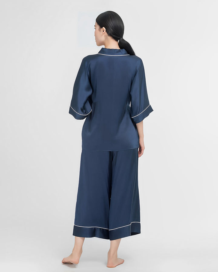 A woman wearing the 19 Momme Silk Wrap Pajamas Set from SusanSilk, featuring dark blue silk with white trim, stands barefoot and faces away on a plain white background.