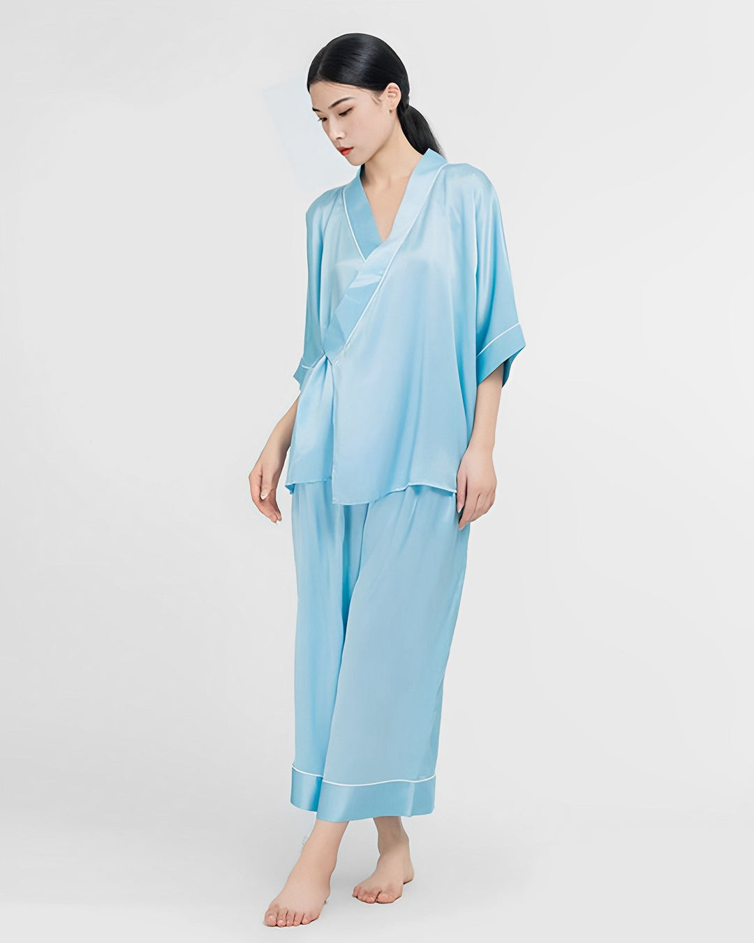 A person wearing SusanSilk's 19 Momme Silk Wrap Pajamas Set in light blue stands against a white background.