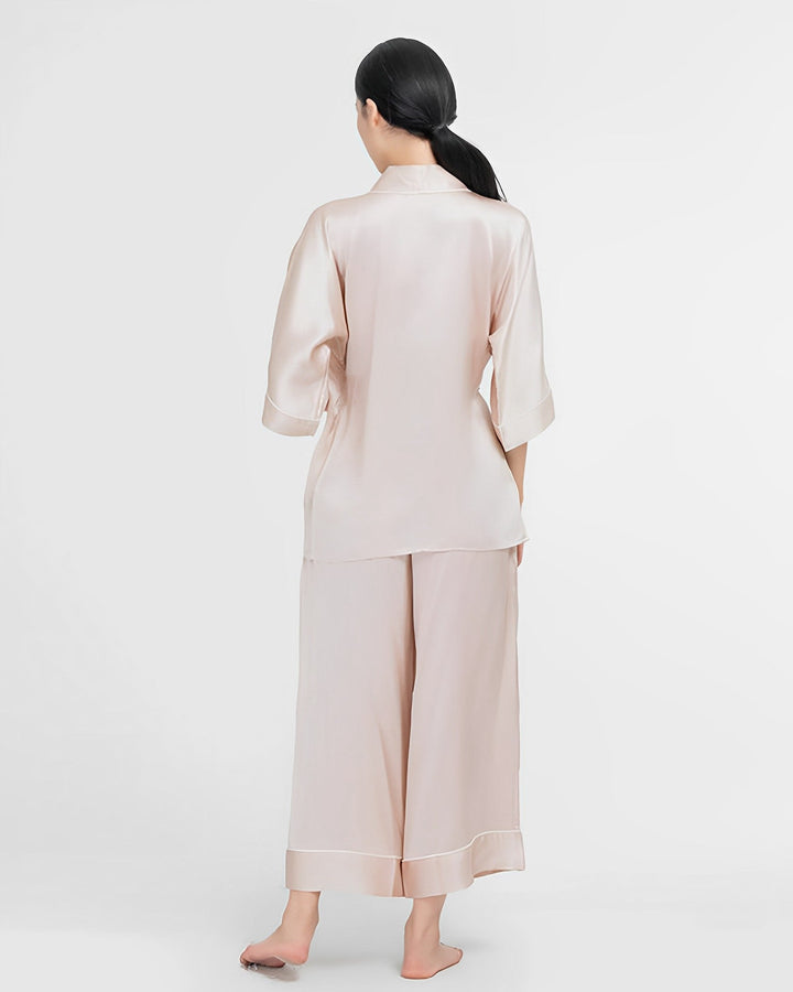 A person with long dark hair, seen from behind, is wearing a light-colored, loose-fitting 19 Momme Silk Wrap Pajamas Set by SusanSilk, featuring three-quarter sleeves and cropped pants, standing barefoot on a white background.