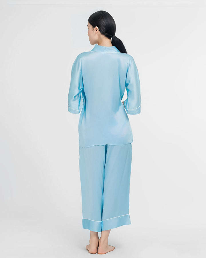 A woman with long, dark hair stands barefoot and faces away, dressed in light blue, loose-fitting silk pajamas from the 19 Momme Silk Wrap Pajamas Set by SusanSilk.