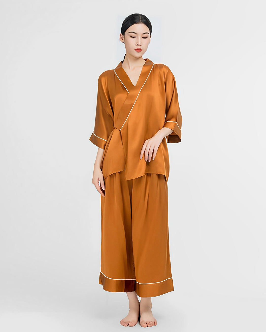 A person stands barefoot sporting the SusanSilk 19 Momme Silk Wrap Pajamas Set, featuring loose-fitting three-quarter sleeves and wide-legged pants in a beautiful burnt orange color, against a plain white background.