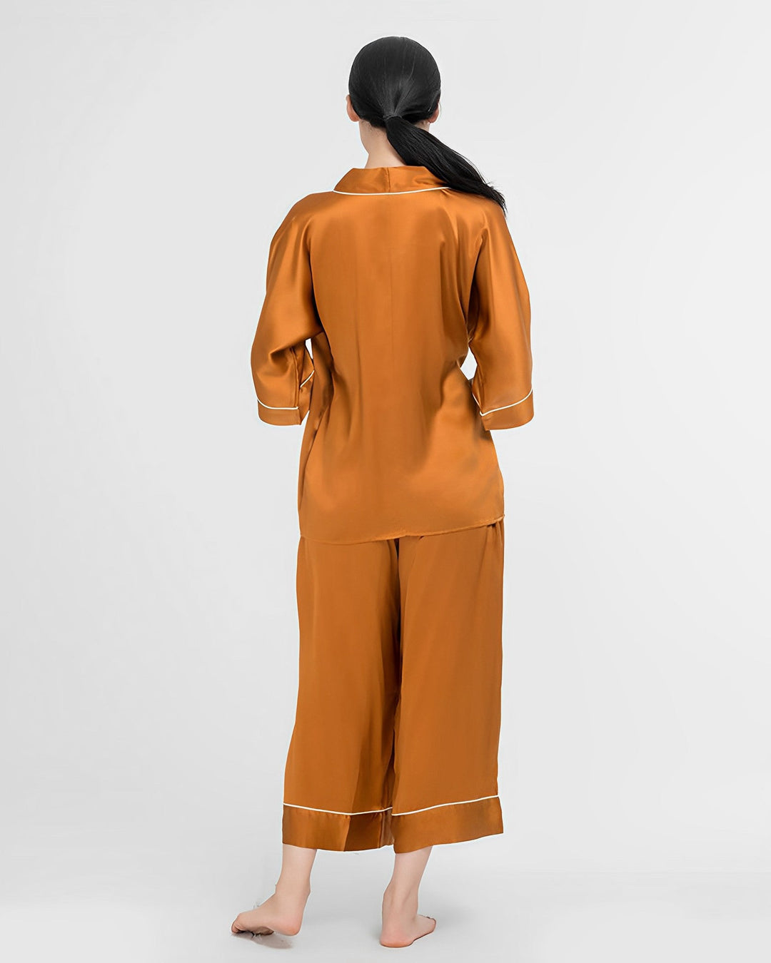 A person with long dark hair is standing barefoot and facing away from the camera, wearing a SusanSilk 19 Momme Silk Wrap Pajamas Set in orange, featuring long sleeves and white trim.