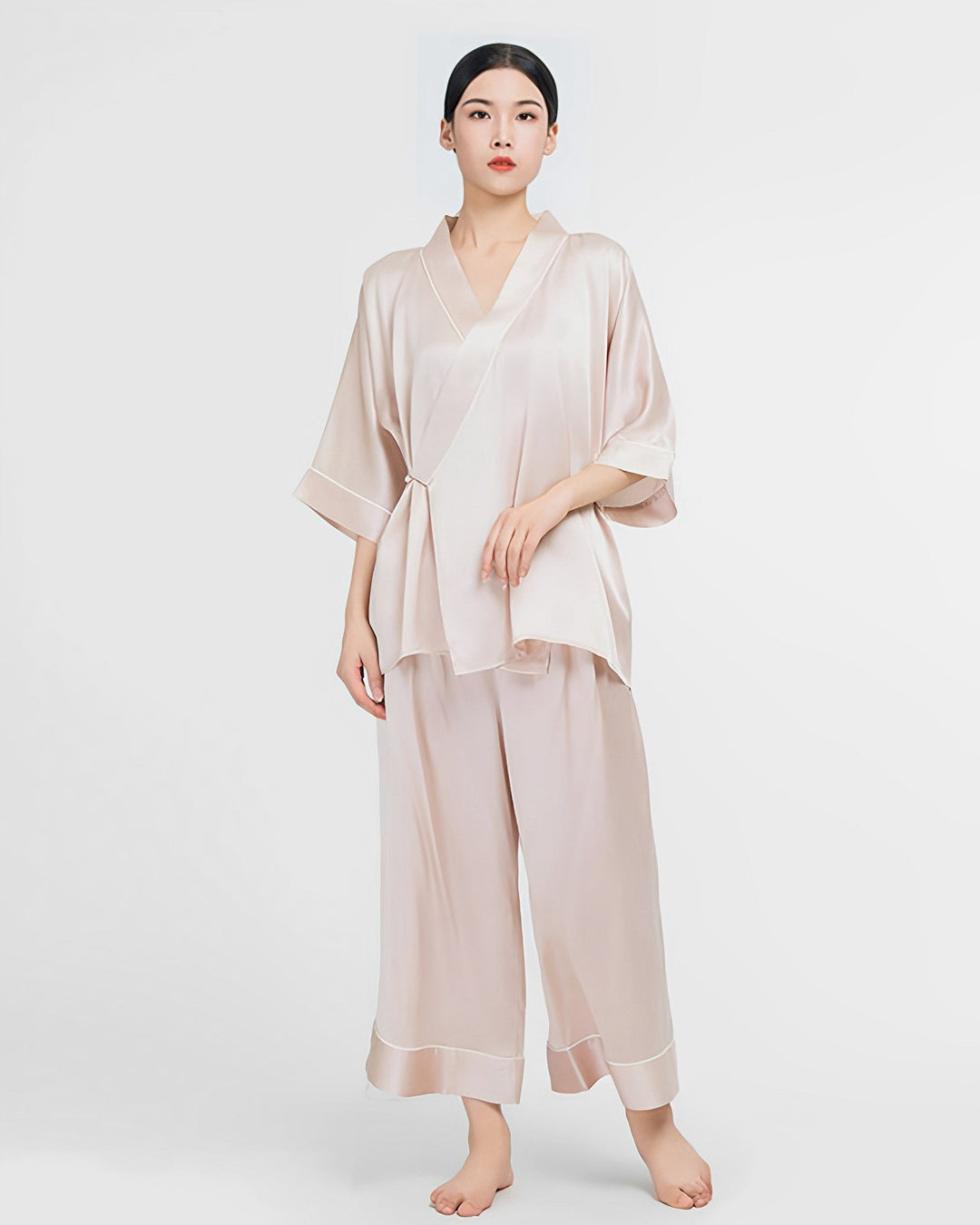 A person stands against a white background wearing a beige, long-sleeve, loose-fitting SusanSilk 19 Momme Silk Wrap Pajamas Set with matching wide-legged pants.