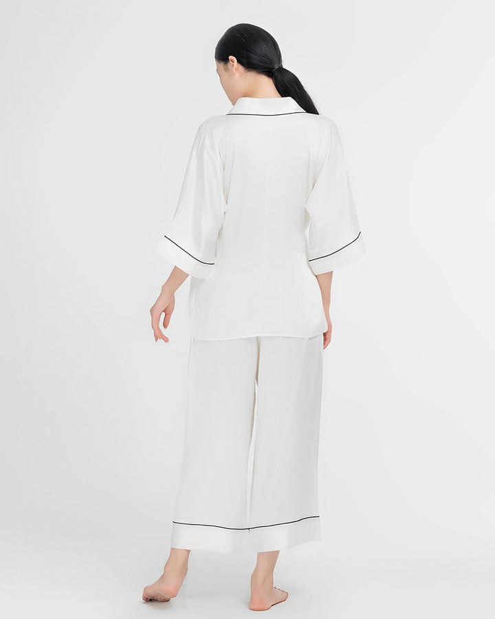 A person standing barefoot in the SusanSilk 19 Momme Silk Wrap Pajamas Set, featuring white silk with black trim, facing away.