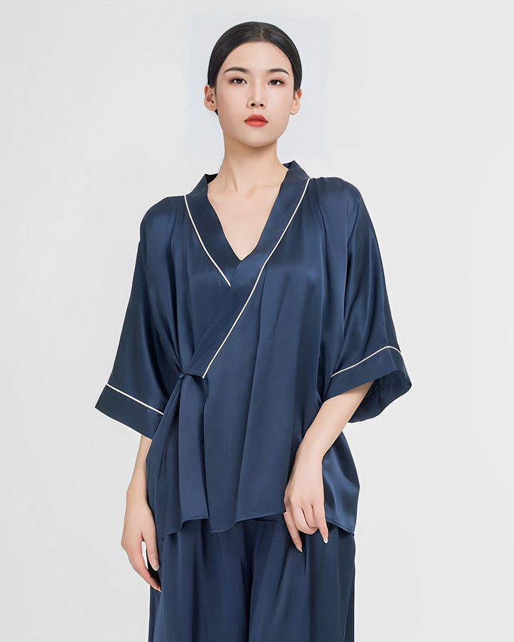 A person wearing a dark blue 19 Momme Silk Wrap Pajamas Set from SusanSilk stands against a plain light background, with a relaxed and confident expression. The outfit features a V-neck top with three-quarter sleeves and matching pants.