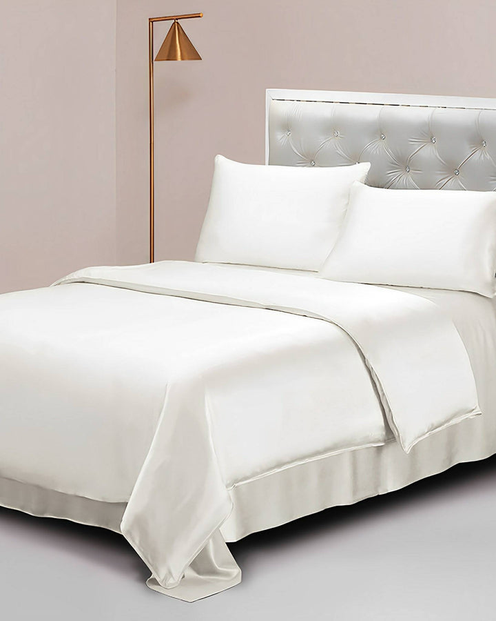 A neatly made bed with a white SusanSilk 19 Momme Seamless Silk Duvet Cover, silk pillows, and a quilted headboard. A gold floor lamp is positioned beside the bed.