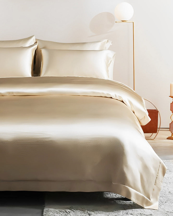 A neatly made bed with a light cream-colored 19 Momme Seamless Silk Duvet Cover with Flange Border from SusanSilk, along with matching pillows. A floor lamp with a round globe and a side table are in the background.