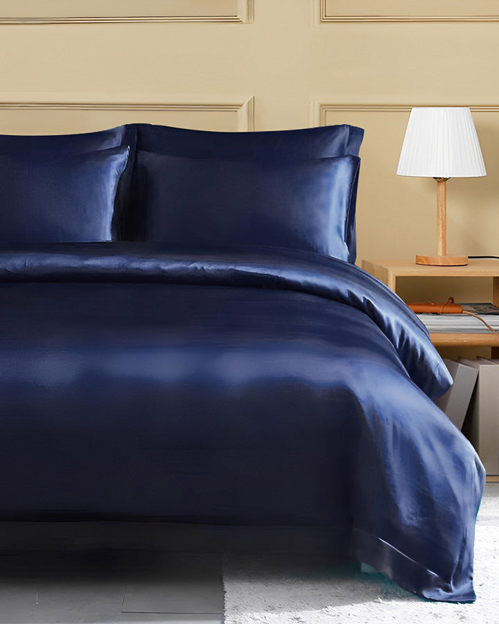 A bedroom with a neatly made bed featuring luxurious SusanSilk's 19 Momme Seamless Silk Duvet Cover in dark blue, complete with matching pillowcases. Beside the bed stands a wooden nightstand topped with a white lampshade. Beige paneled walls form the backdrop.