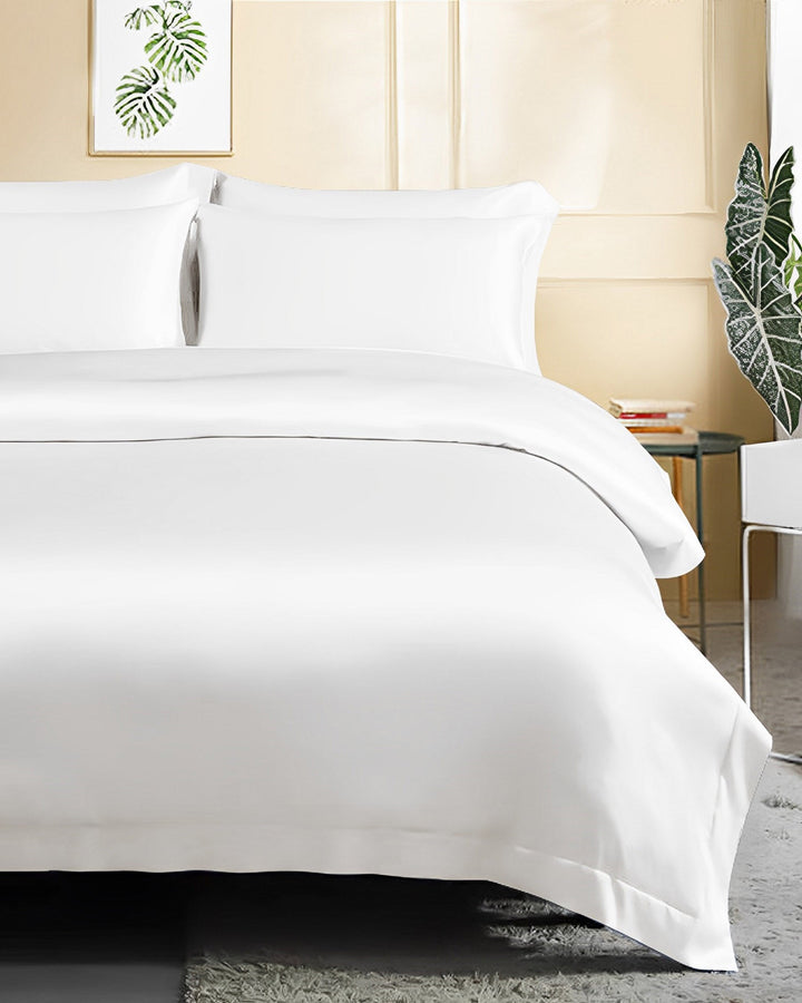 A neatly made bed adorned with SusanSilk's 19 Momme Seamless Silk Duvet Cover - Flange Border, accompanied by white sheets and plush pillows. The room showcases light-colored walls, vibrant green plants in the background, and a sleek side table with a glass top.