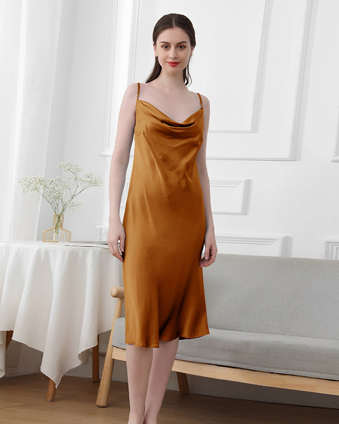 A woman dressed in an Elegant Cowl Neck Silk Nightdress by SusanSilk stands in a minimalist room with white walls. A vase with white flowers from SusanSilk is on a table beside her.